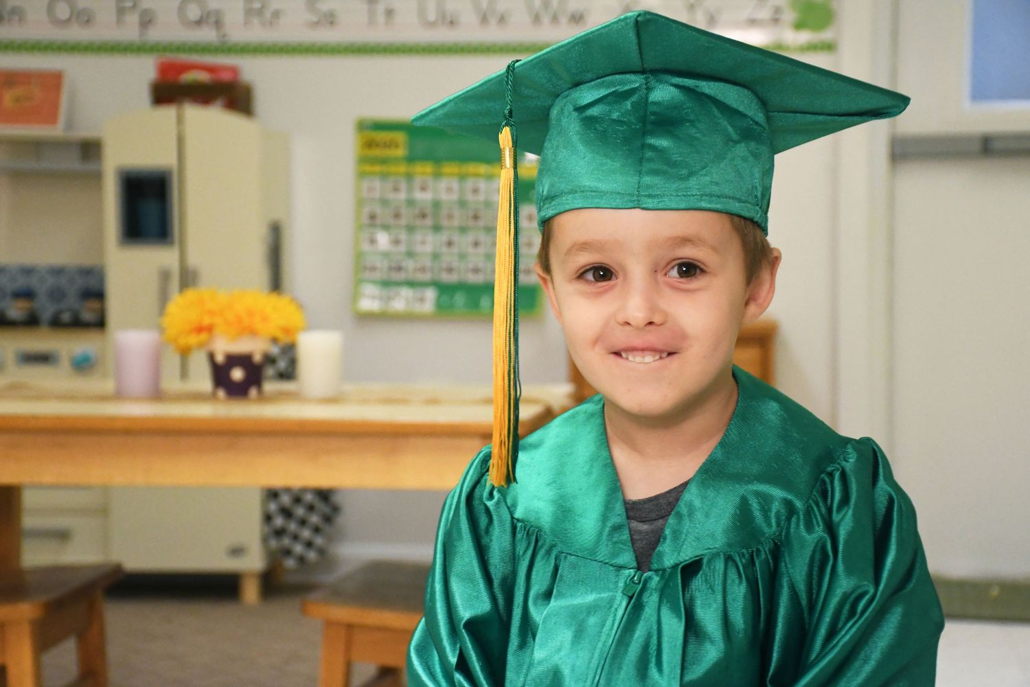Green-robed graduates pose together in an elementary school classroom setting.