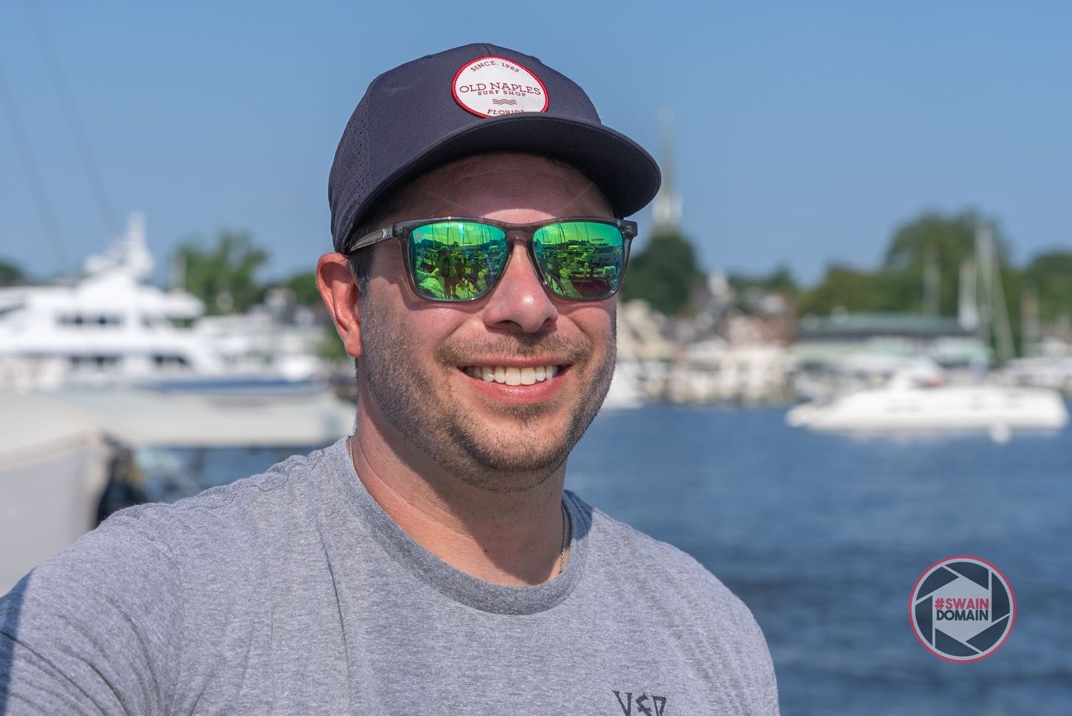 Someone wearing sunglasses with green reflective lenses and a baseball cap poses at a marina with boats in background.