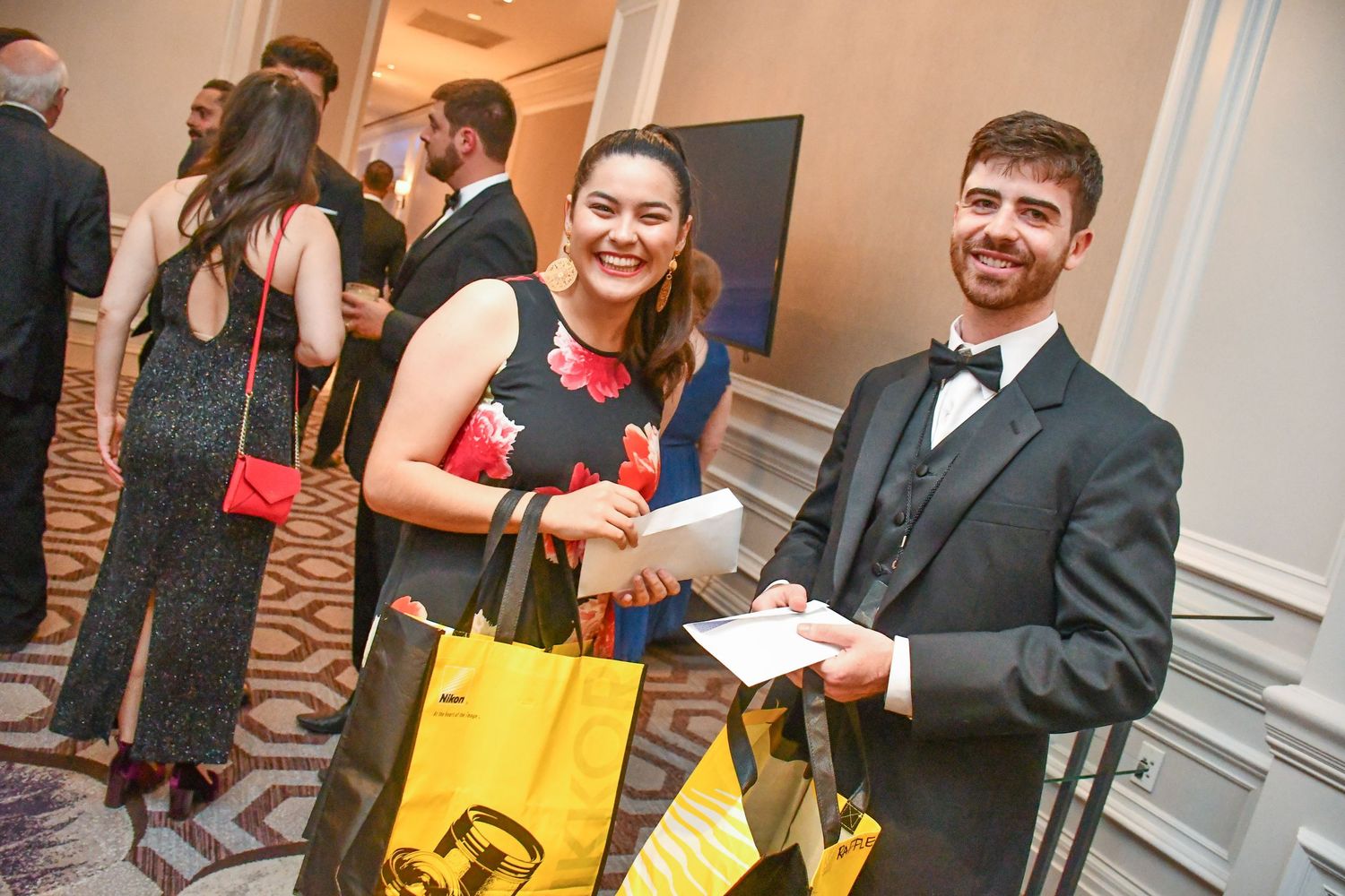 Two formally dressed guests smile at a gala event, holding yellow gift bags and papers in an elegant ballroom.