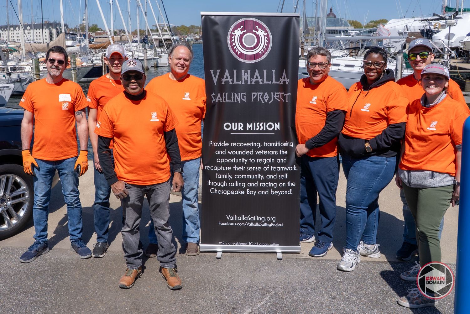 Group of volunteers in bright orange t-shirts stand by Valhalla Sailing Project banner at marina dock.