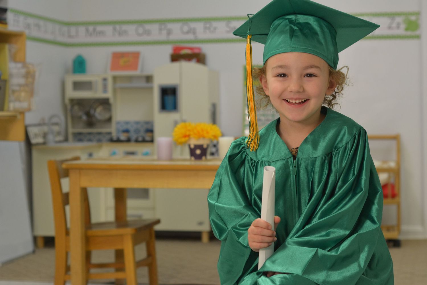 Students in emerald graduation attire gather in a classroom with alphabet decorations.