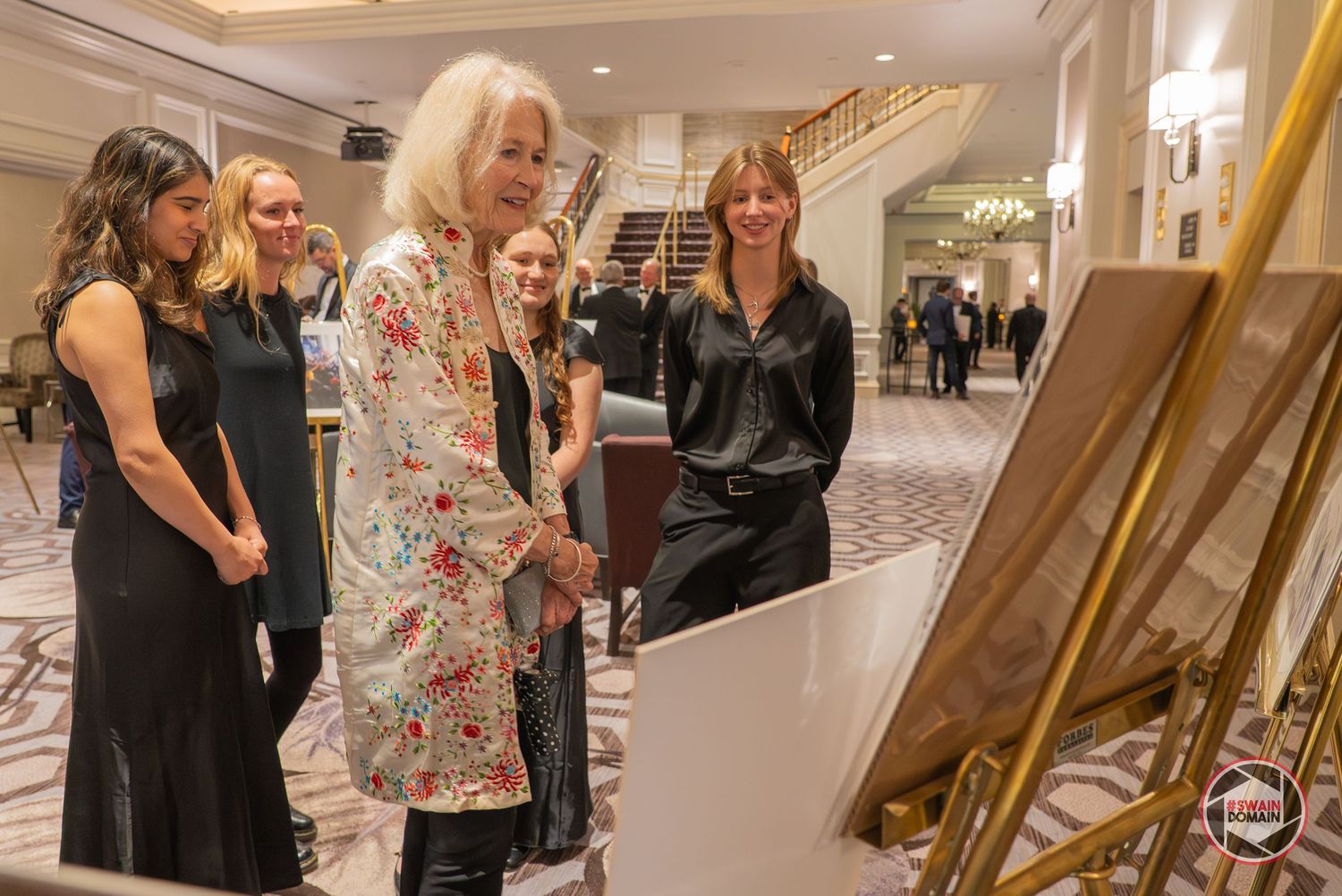 Elegantly dressed women viewing artwork on an easel at a formal gala event in a grand hotel ballroom.