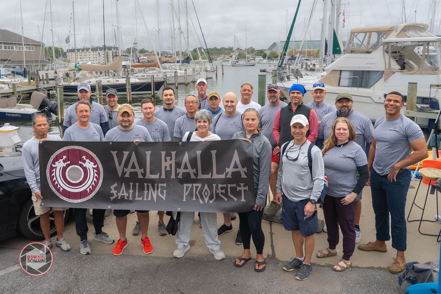 A group of people gathered outdoors near boats and a sailing club sign in a marina setting.