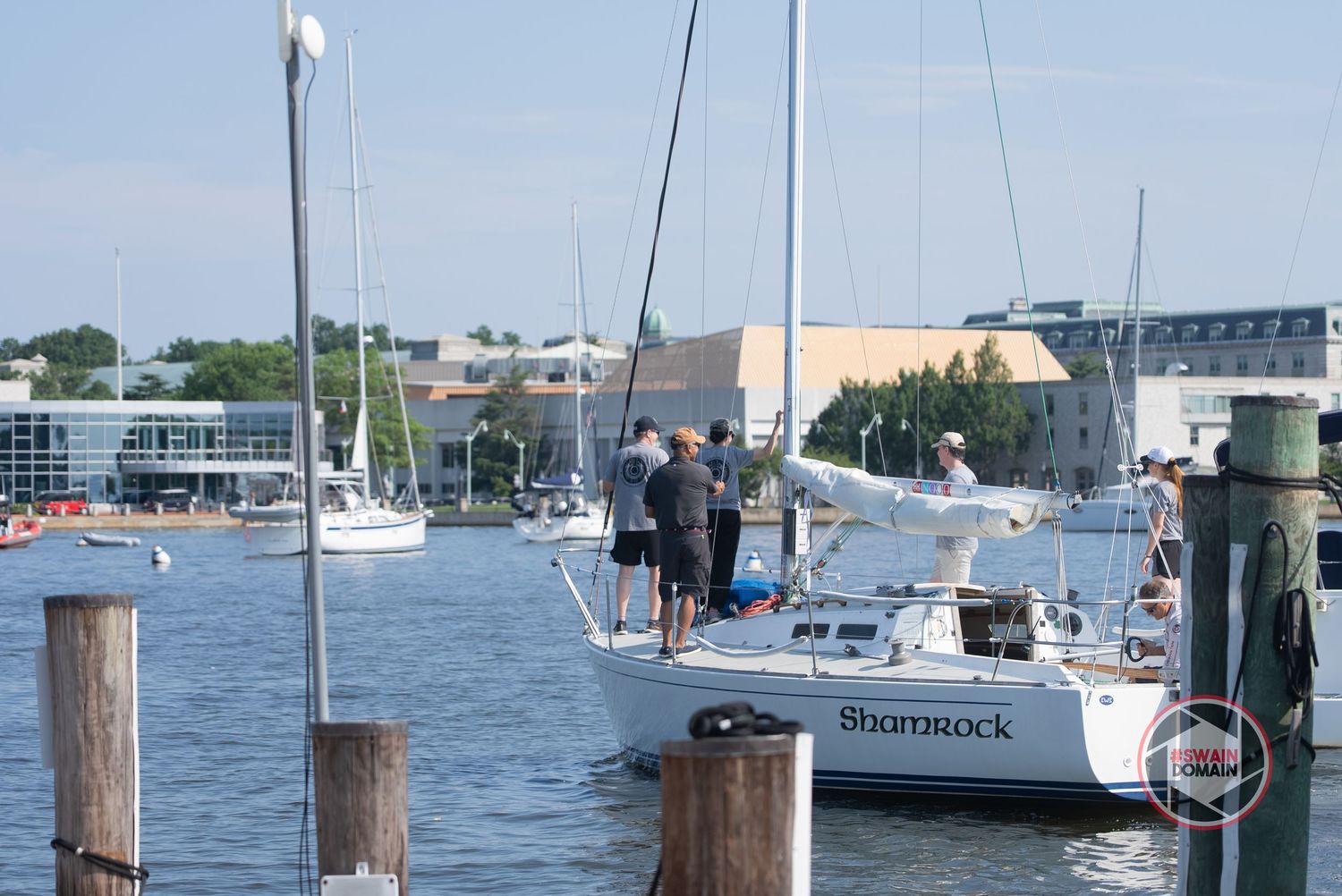 People standing on a sailboat docked at a marina with other boats and buildings visible in the background.