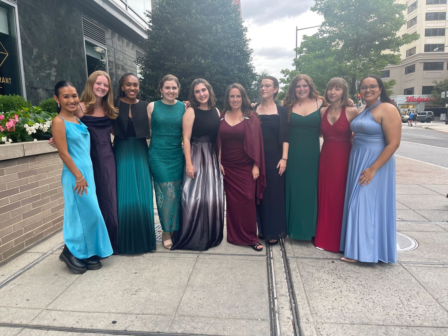 Group of women in elegant formal gowns posing together outdoors on a sidewalk at a special event.