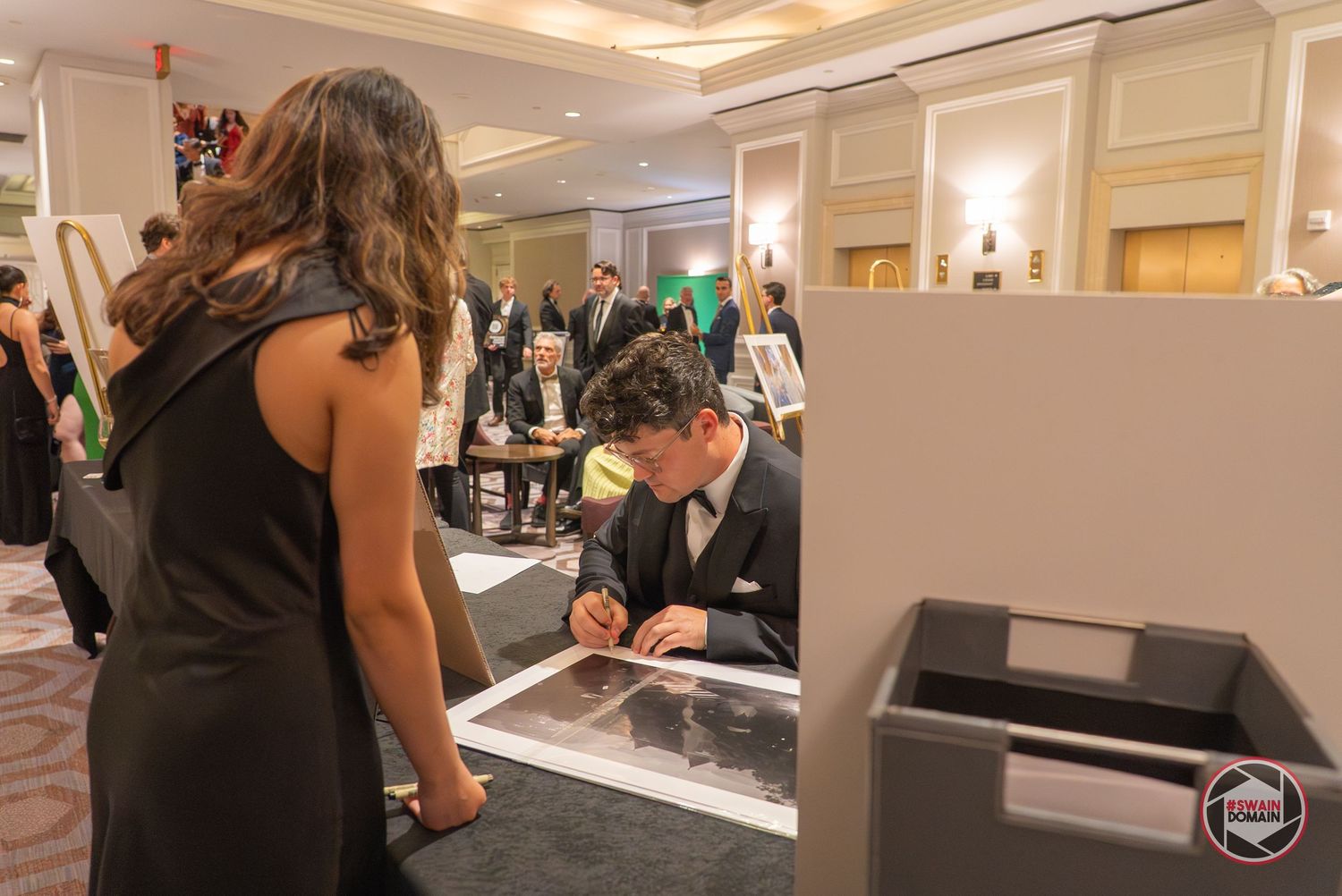 Man in tuxedo signing artwork at elegant gala event while guests observe in formal ballroom setting.