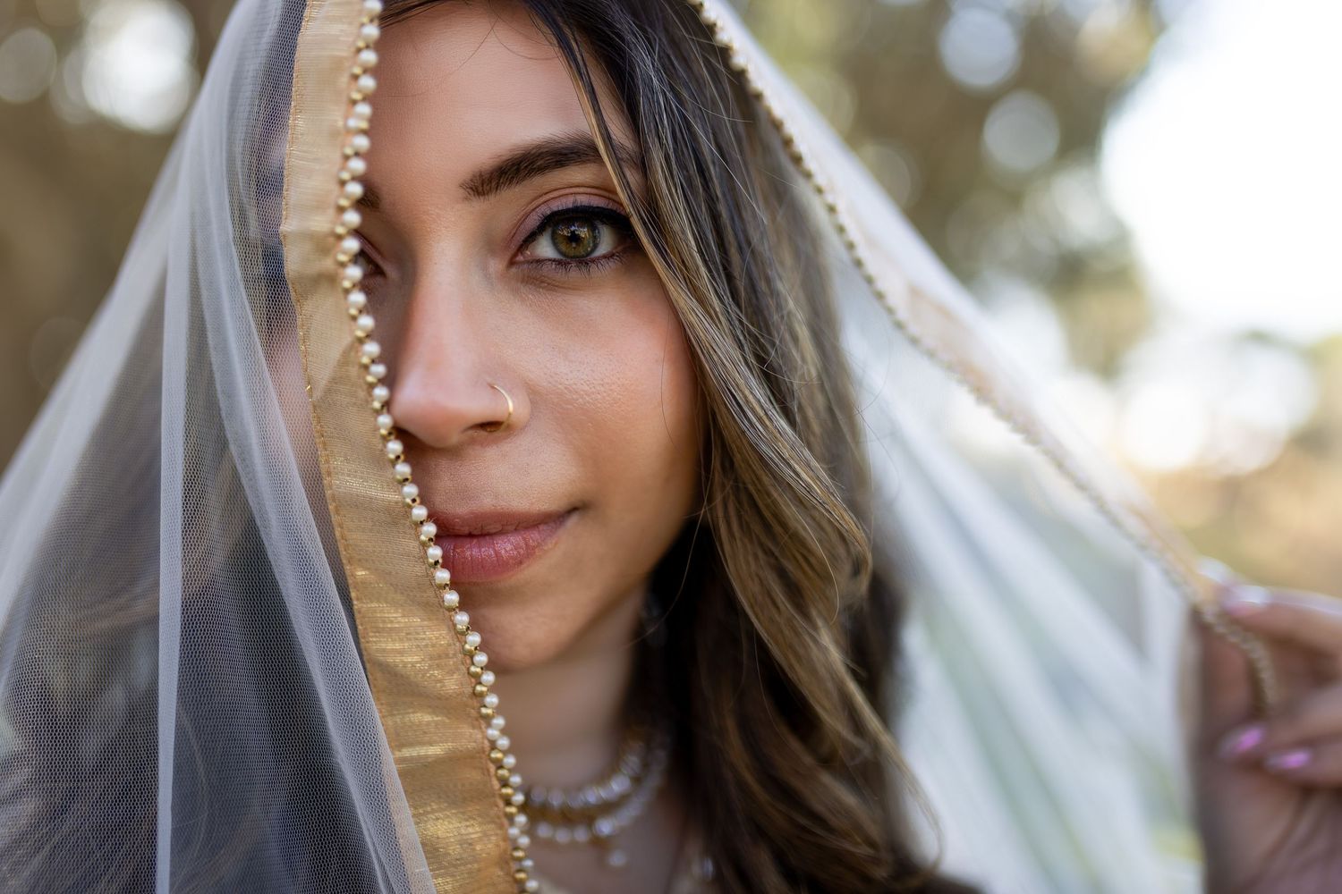 Bride wearing a pearl-trimmed veil draped over her face, showcasing elegant bridal jewelry in an outdoor setting.