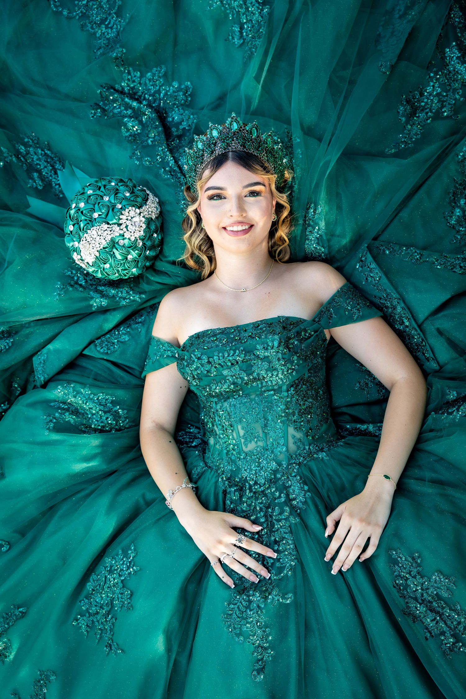 Young woman in emerald green off-shoulder quinceañera ball gown lying down, wearing matching crown and holding floral bouquet.