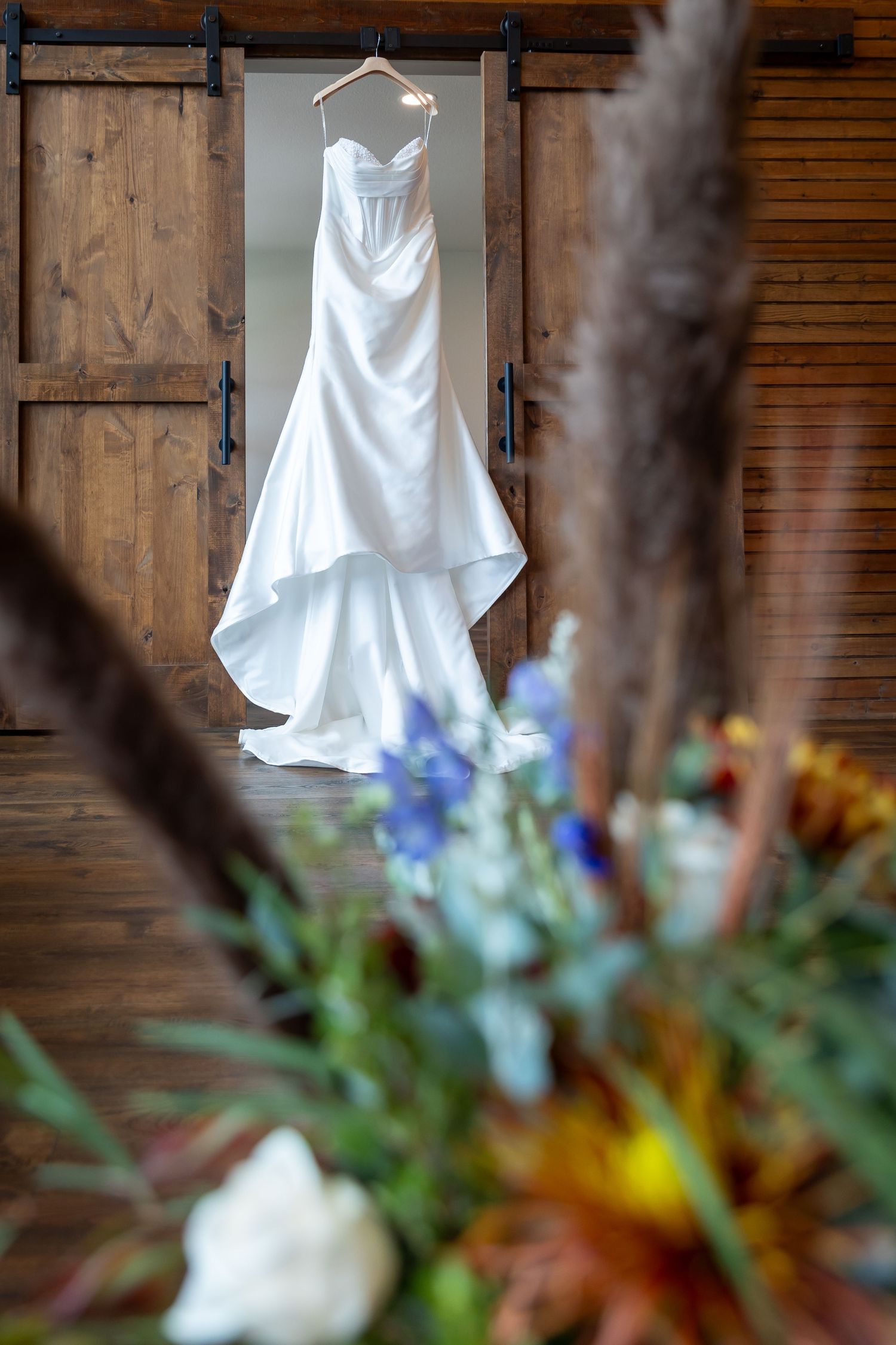 White satin mermaid wedding dress hanging in a rustic barn doorway, framed by blurred blue and orange wildflowers and pampas grass.