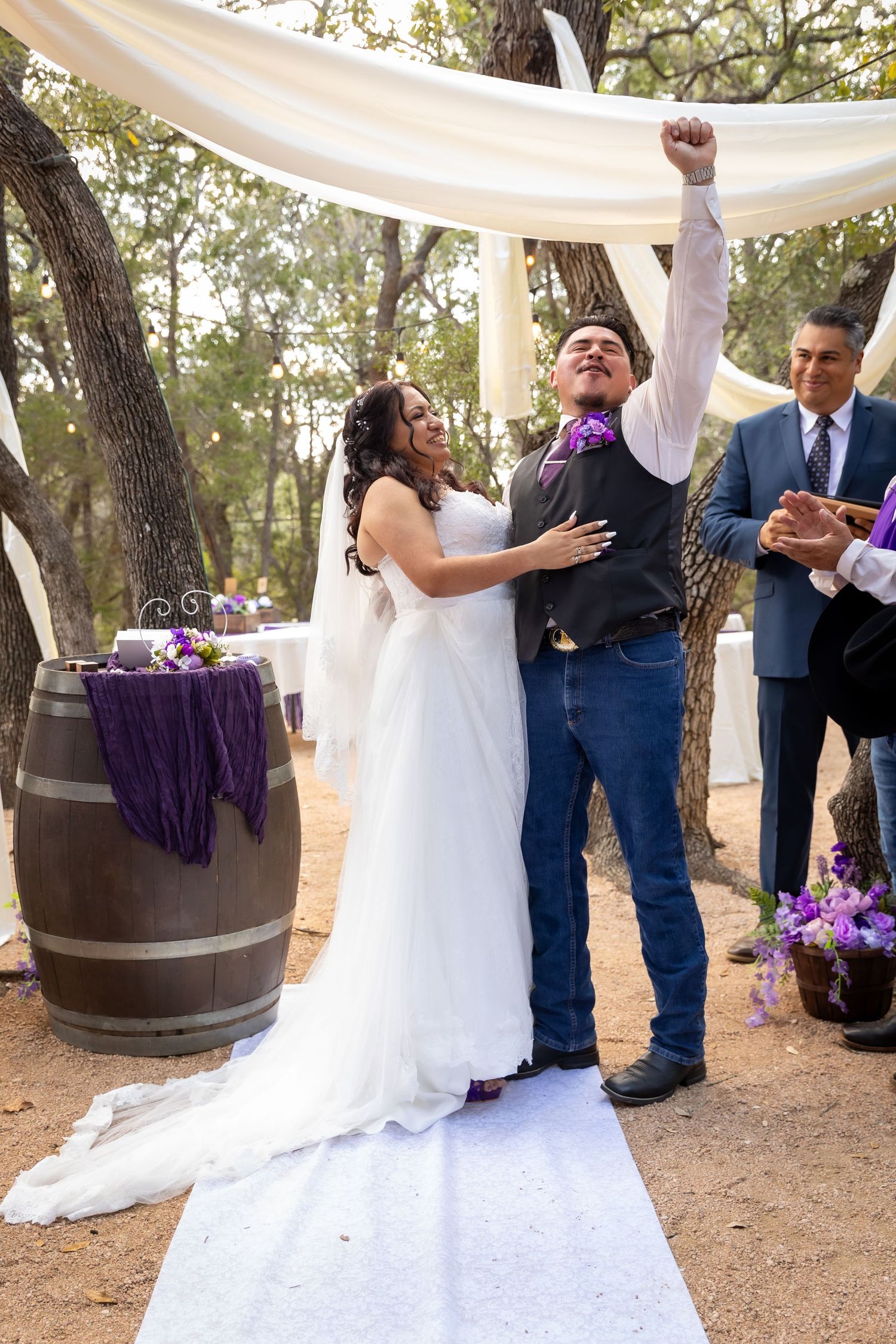 Joyful groom raises his fist in celebration beside his bride in a white gown during an outdoor wedding ceremony with purple accents.
