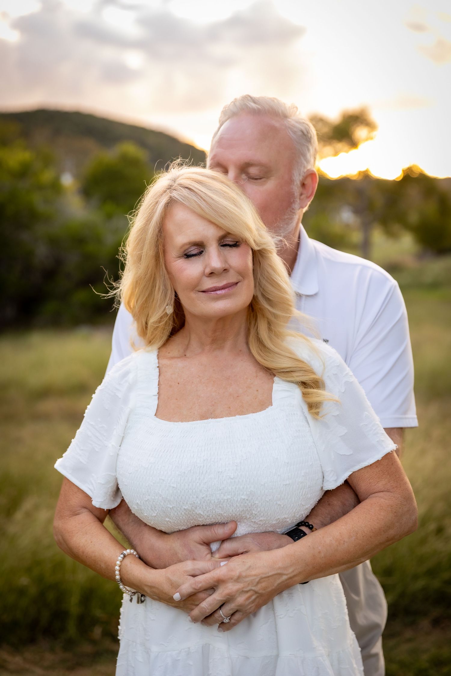 Blonde woman in a white dress closes her eyes as a gray-haired man embraces her from behind in a golden-hour outdoor couple portrait.