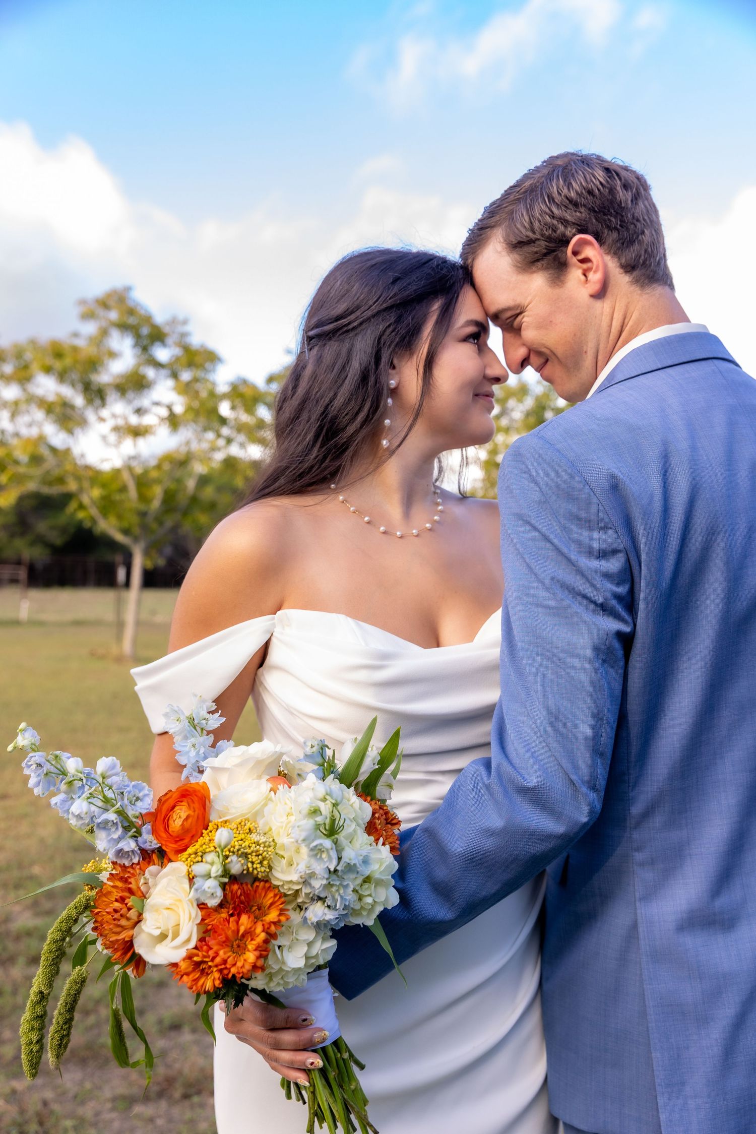 Bride in an off-shoulder white gown holding an orange and white bouquet touches foreheads with her groom in a blue suit outdoors.