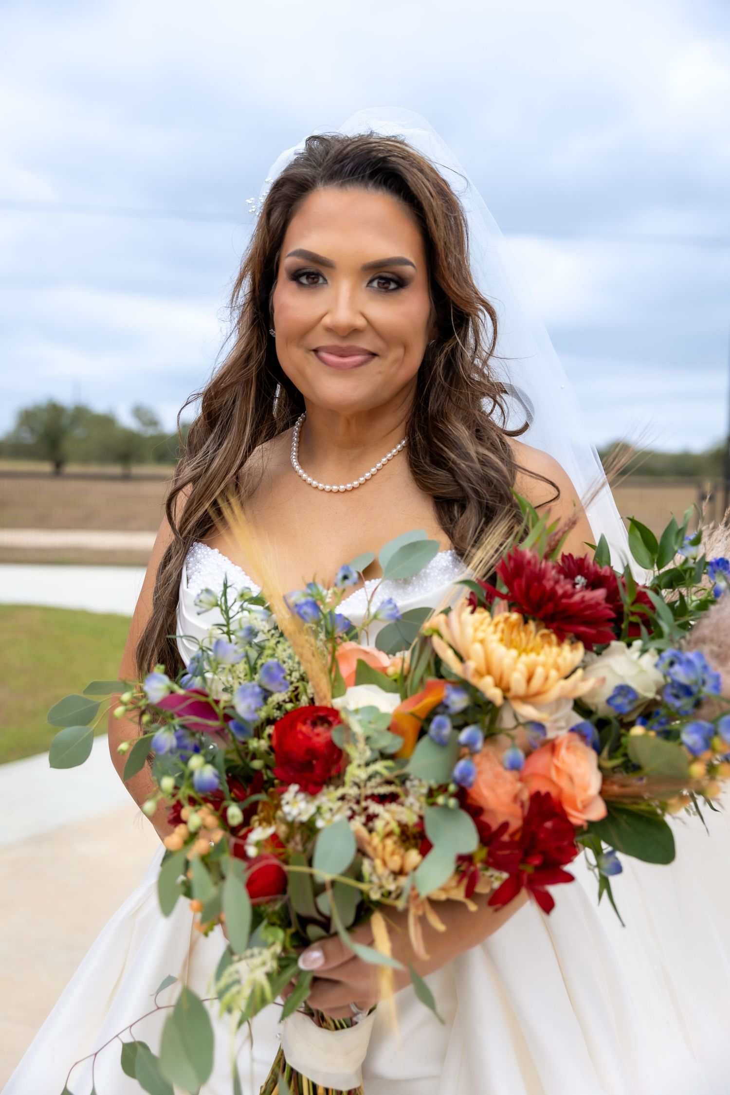 Smiling bride in a white gown, veil and pearl necklace holds a colorful bouquet of red, orange and blue flowers outdoors.