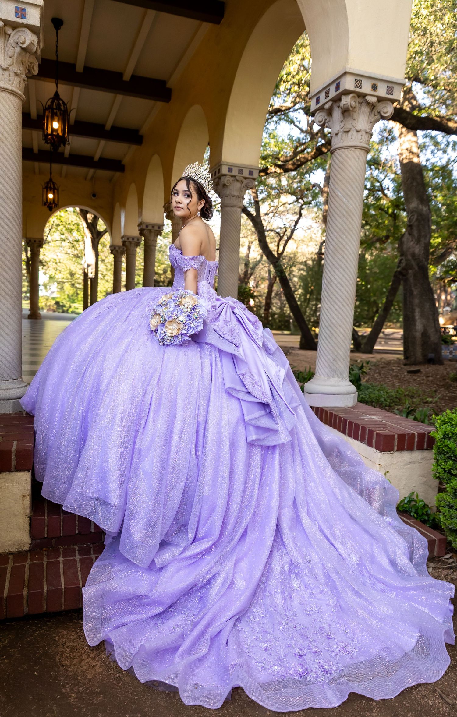 Young woman in a lavender ball gown with floral bouquet poses gracefully under arched columns for quinceañera.