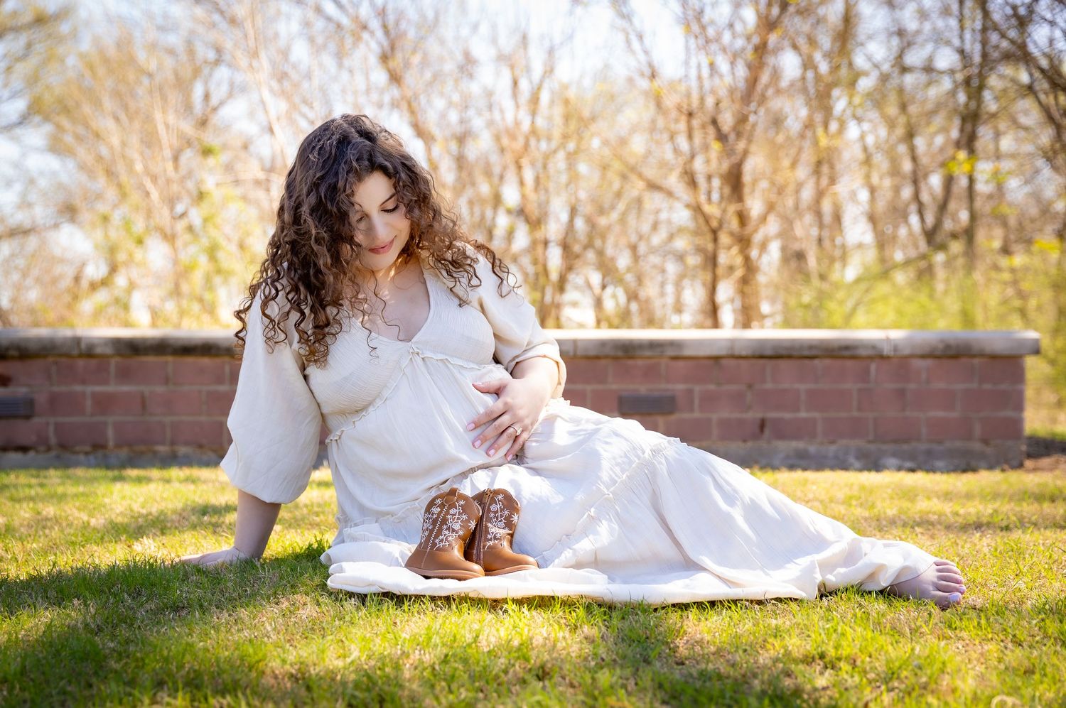 Pregnant woman in white dress sitting on grass, cradling baby bump with tiny boots nearby, outdoor maternity photo.