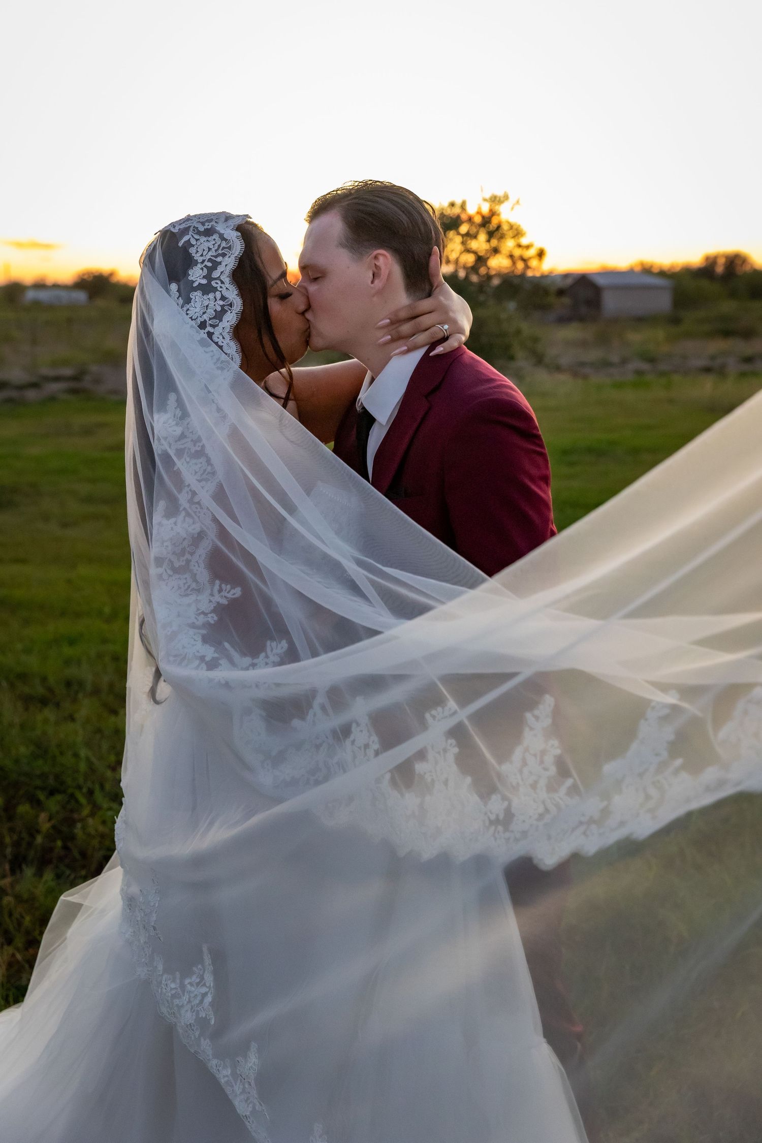 Bride in a lace wedding gown and flowing veil kisses her groom in a burgundy suit in a grassy field at golden sunset.