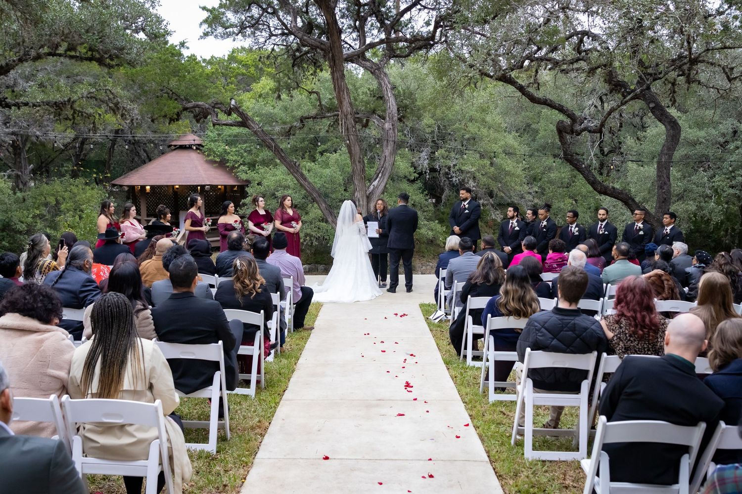 Outdoor wedding ceremony setup with white chairs and rose petals lining the aisle under tall pine trees.