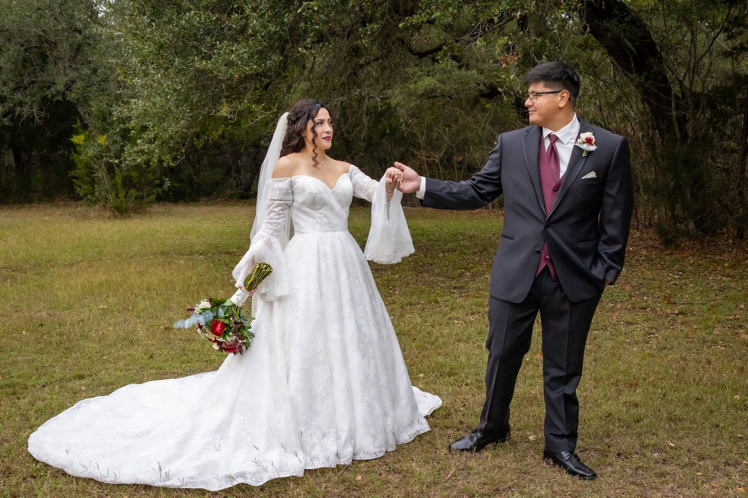 A bride in a lace wedding gown and groom in a dark suit share a moment together on grassy grounds.