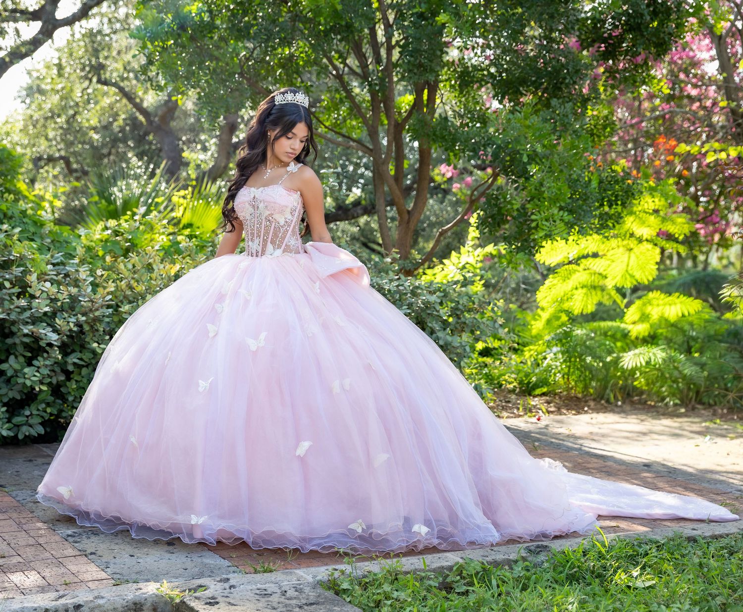 Young woman in a pink ball gown quinceanera dress with floral details poses in a lush garden setting.