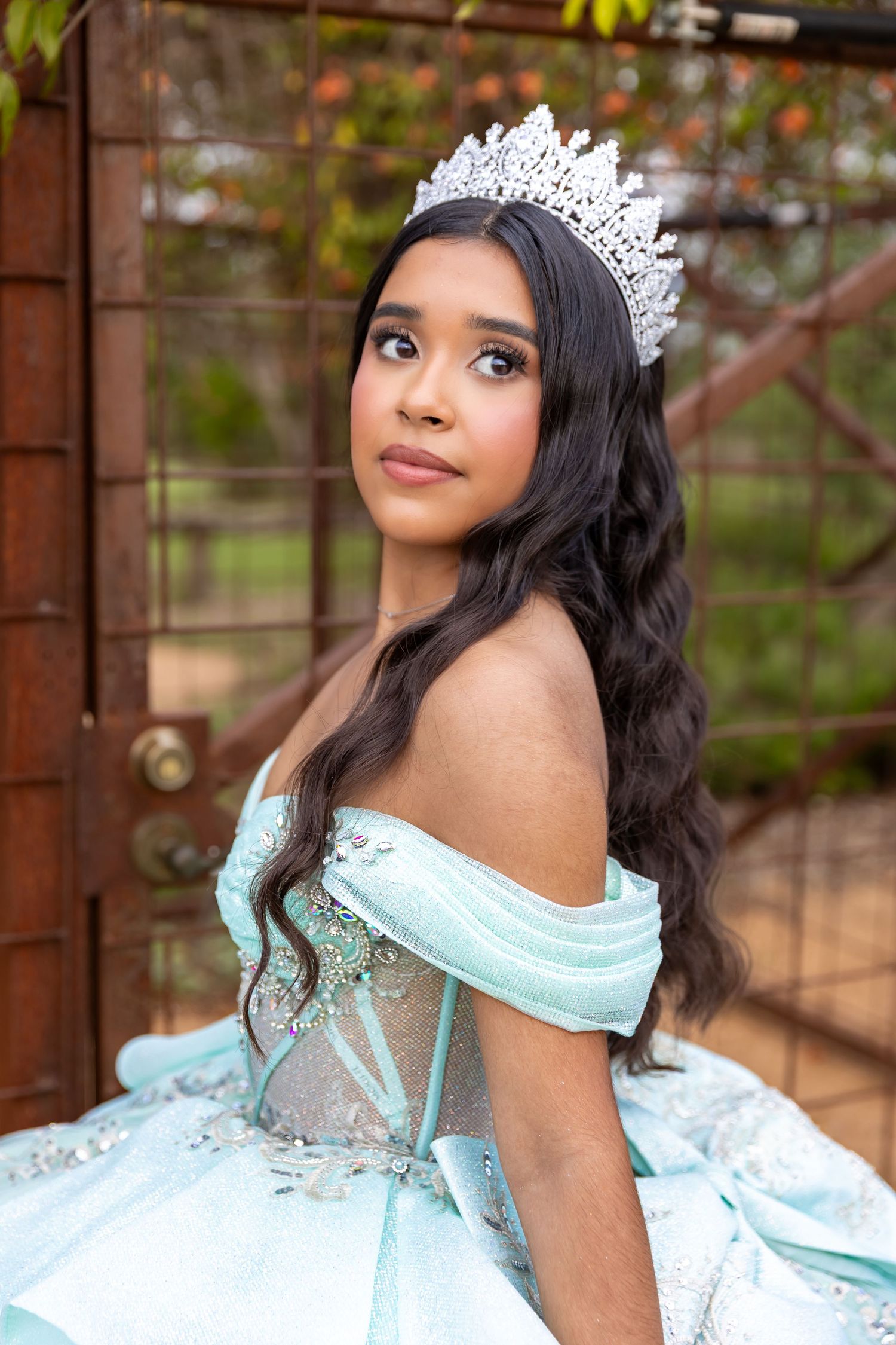 Young woman in a light blue off-shoulder quinceañera gown and sparkling tiara poses gracefully outdoors.