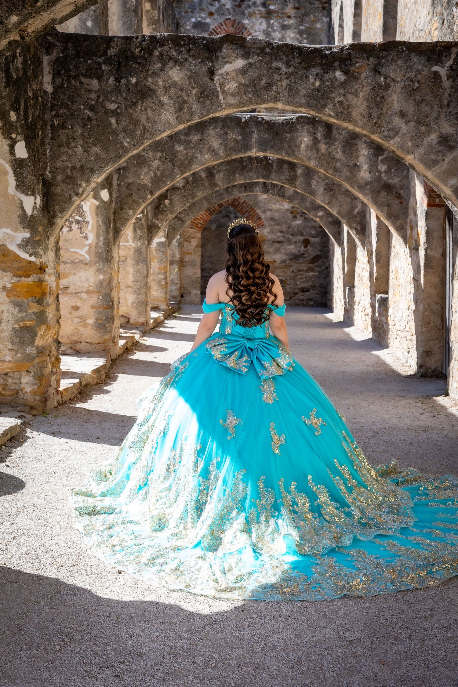 Young woman in turquoise gold quinceañera gown walks through stone archway corridor of historic mission.