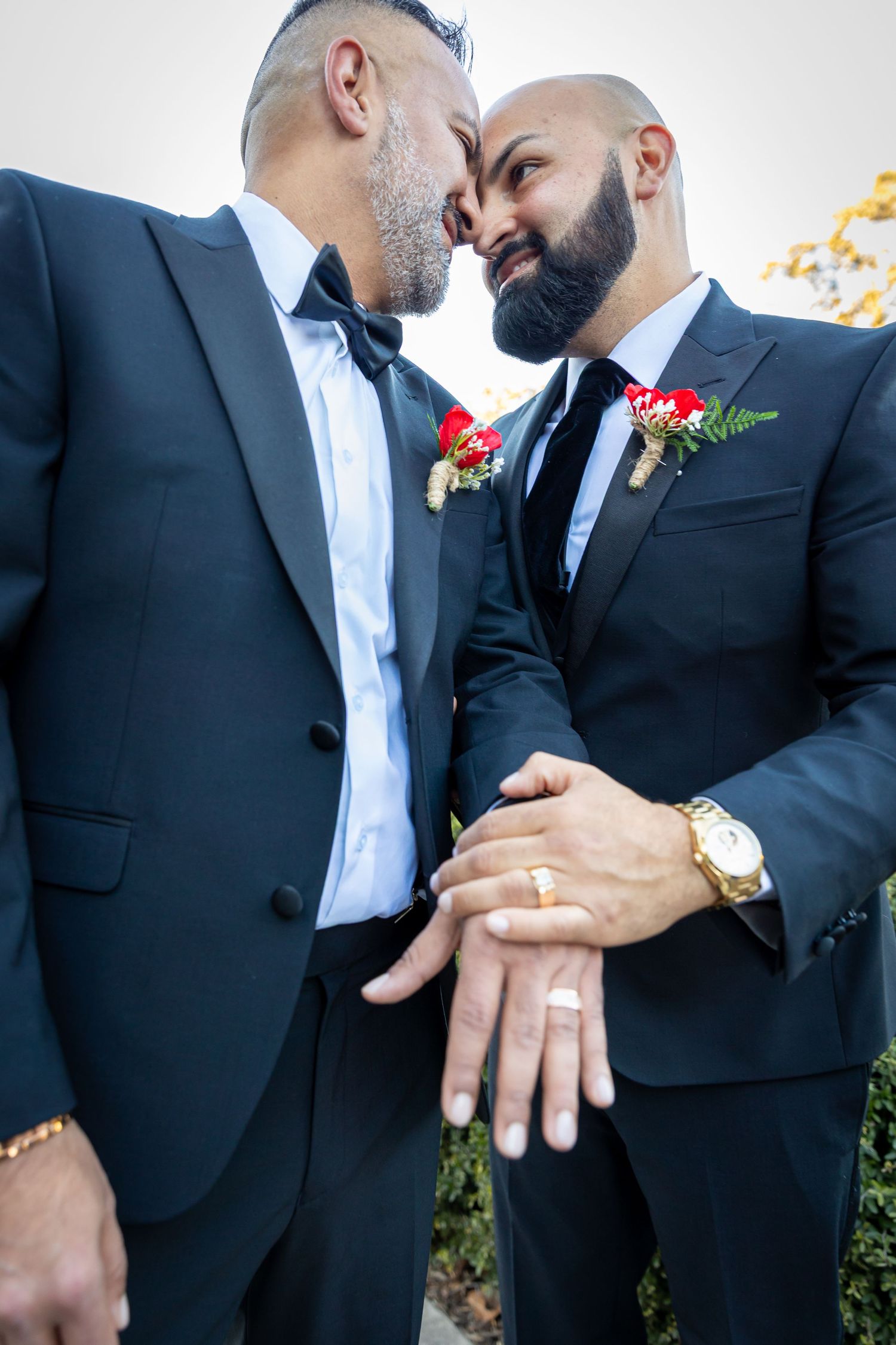 Two grooms in black tuxedos with red boutonnieres share an intimate moment, displaying their wedding rings on their wedding day.