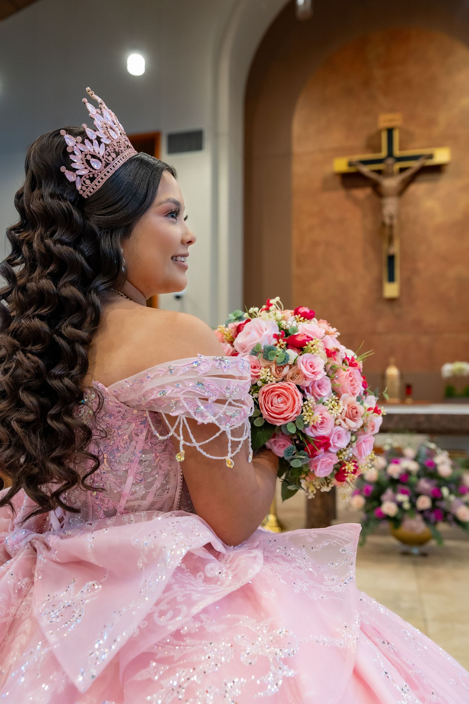 Young Latina in pink quinceañera gown and crown holds floral bouquet inside a church with crucifix backdrop.