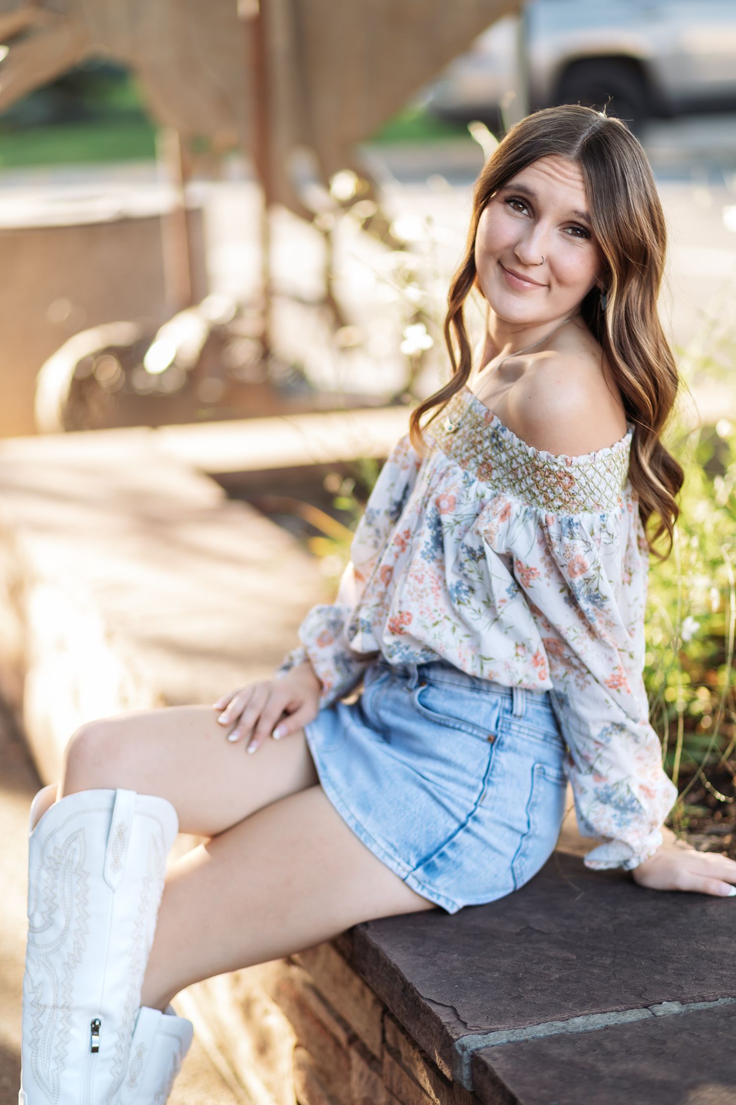 Smiling young woman in a floral off-the-shoulder top, denim skirt and white cowboy boots sits on a stone ledge outdoors.