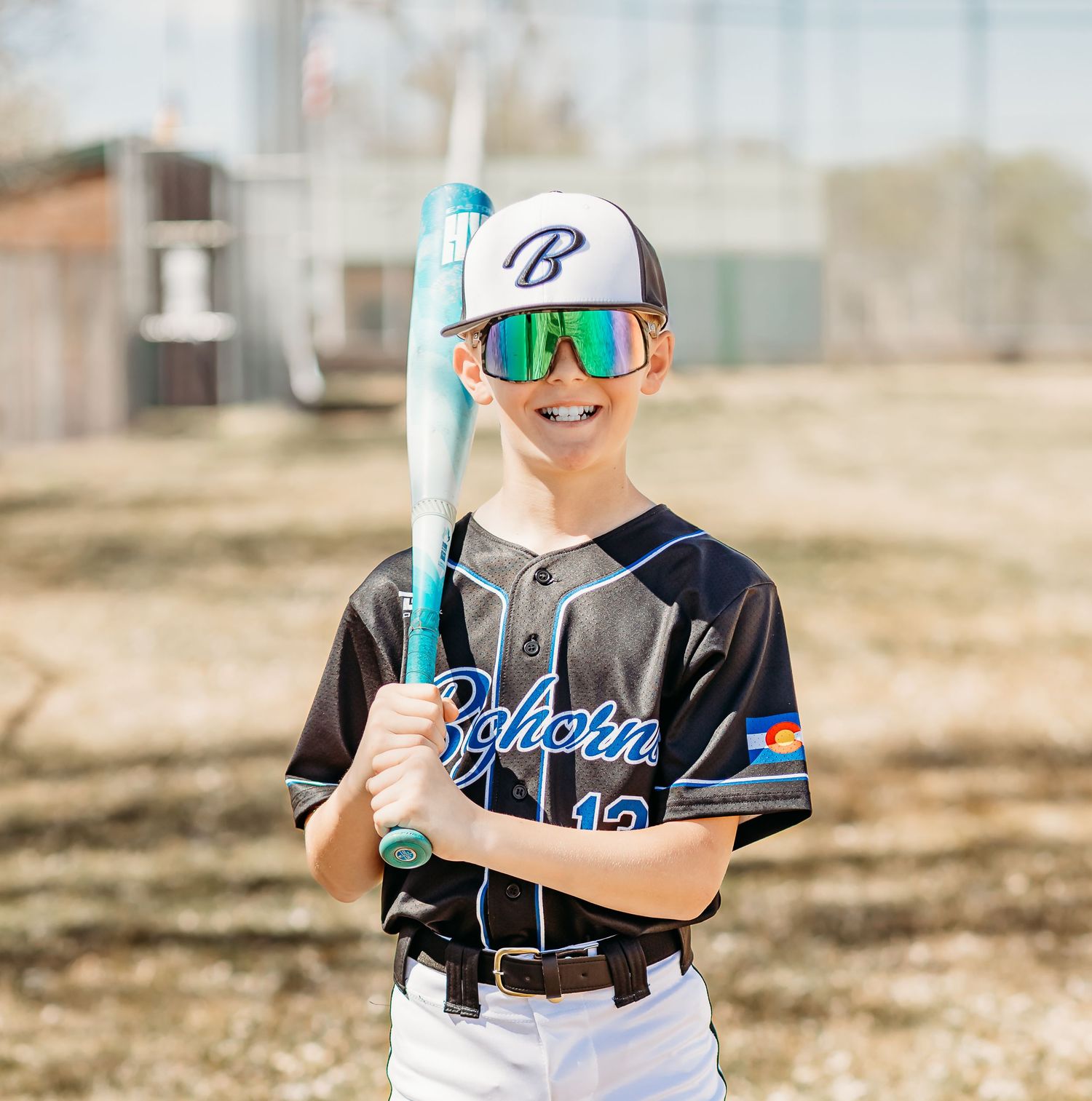 A smiling young baseball player in a black Bighorn jersey, mirrored sunglasses and cap holds a teal bat on a dirt field.