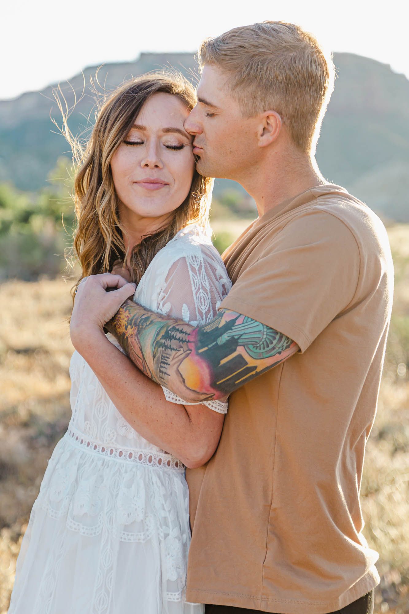 A smiling woman in a white lace dress closes her eyes as a man in a tan shirt kisses her forehead in a sunny desert landscape.