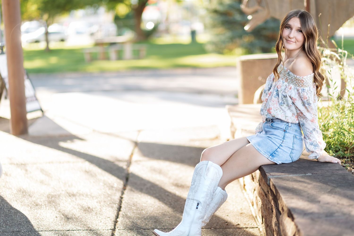 Young woman in a floral off-the-shoulder top, denim shorts and white cowboy boots sits on a stone ledge outdoors.