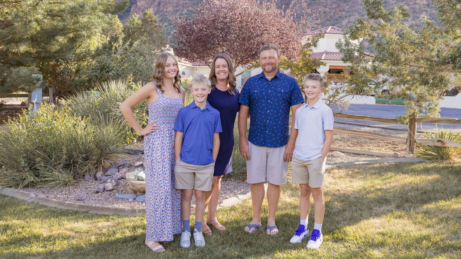 A smiling family of five, two parents and three children, stands together on a grassy lawn surrounded by trees in warm sunlight.