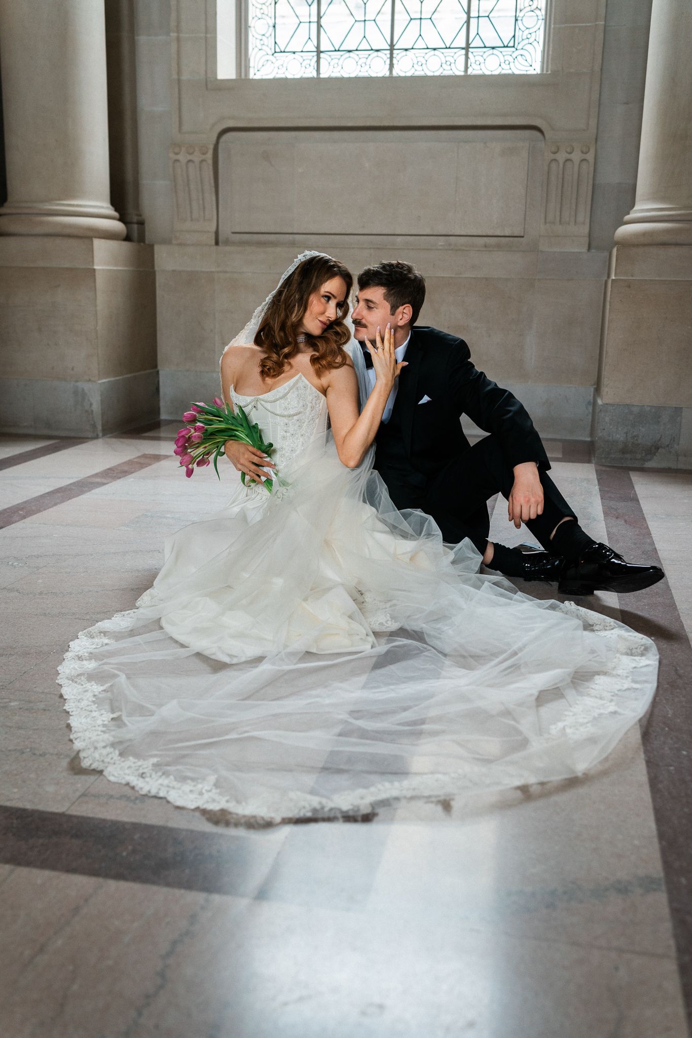 Bride in white lace gown and groom in black suit sit on marble floor, sharing a tender moment with pink bridal bouquet.