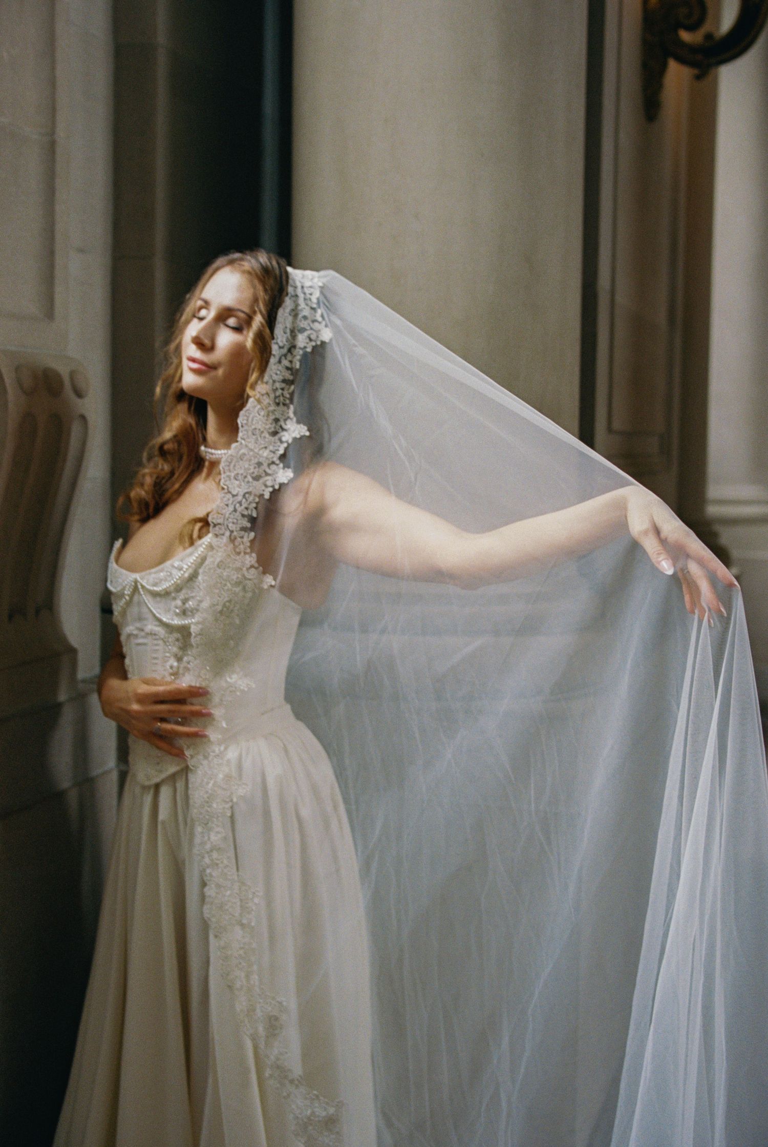 Bride in lace wedding gown holding flowing veil, posing gracefully between stone columns in elegant venue.