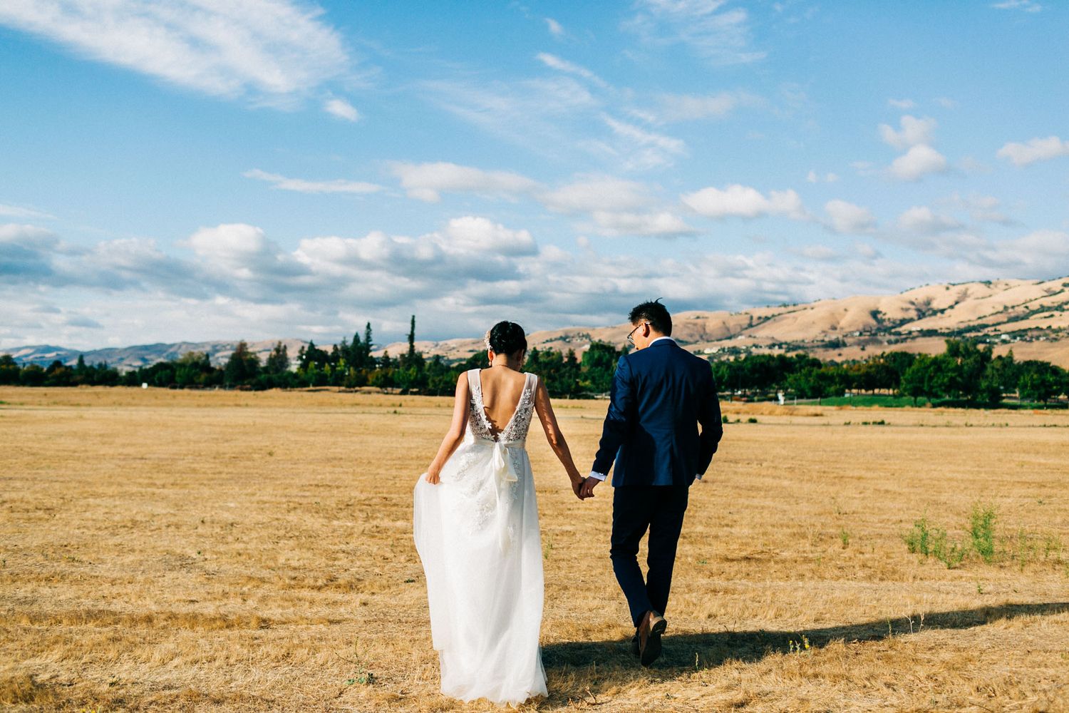 Couple walks through golden wheat fields with mountains and blue sky in the distance on their wedding day.