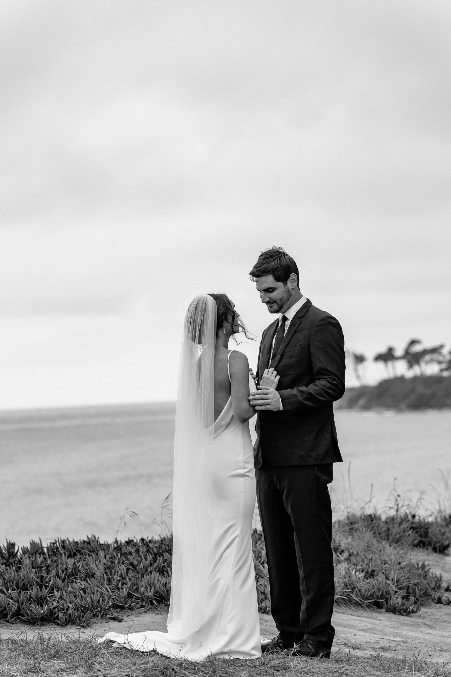 A couple in wedding attire stands together on a coastal cliff overlooking the ocean in black and white photography.