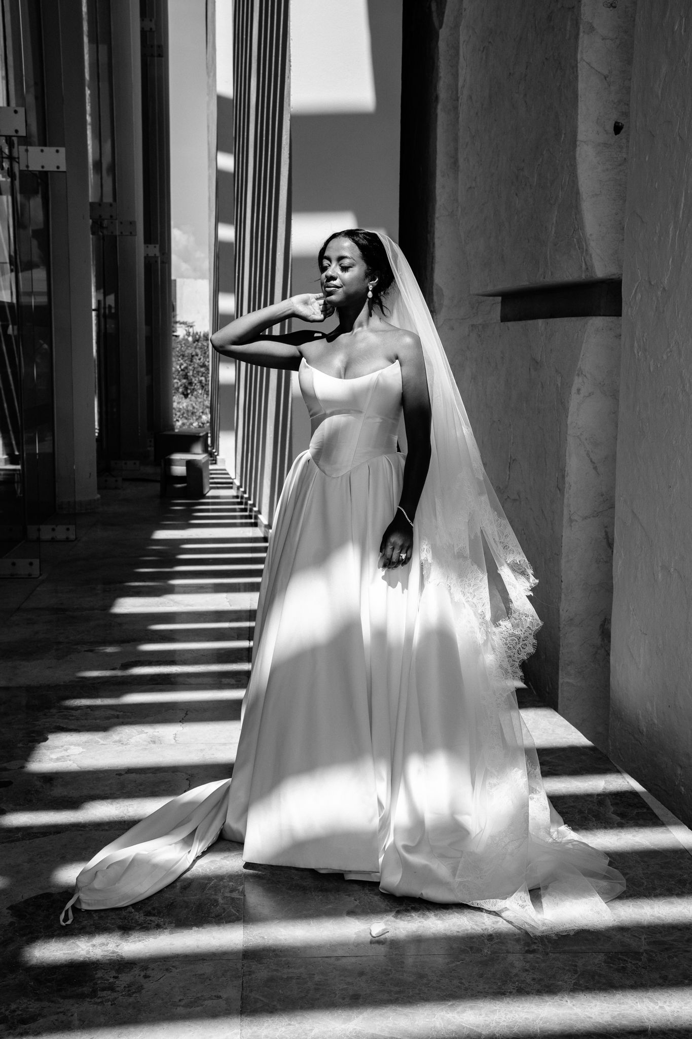 Black and white photo of a bride in a strapless gown and veil posing in dramatic light and shadow.