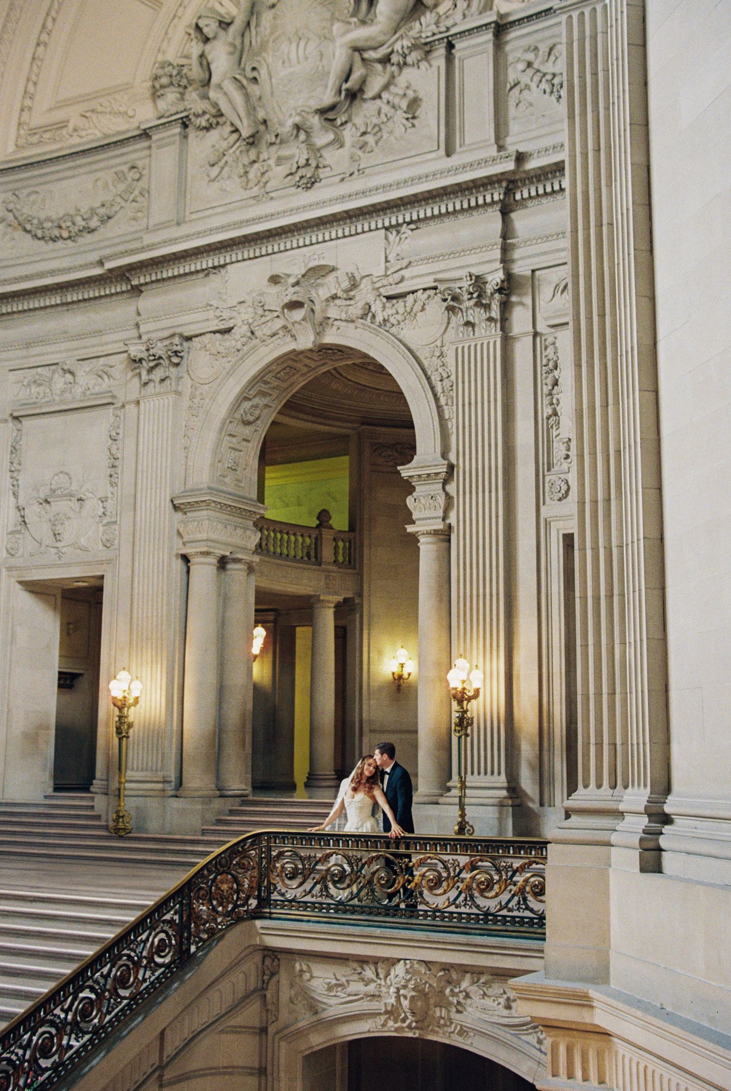 Couple embracing on grand staircase of ornate Beaux-Arts building with elegant columns and arched doorway.