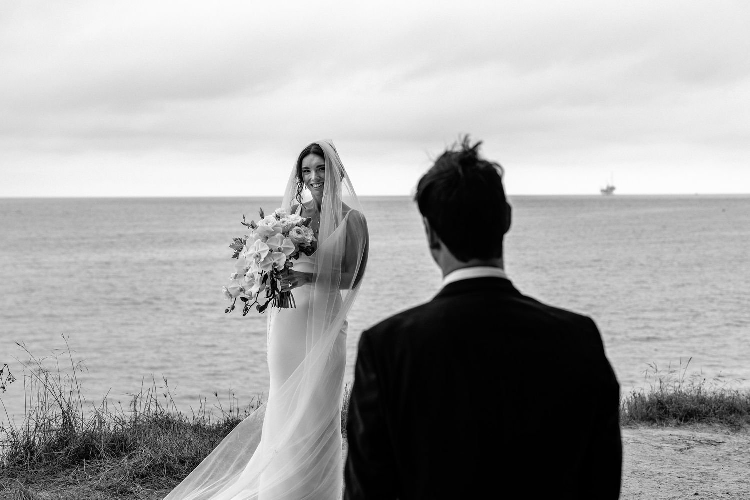 A couple in wedding attire stands against an ocean backdrop as a veil flows in the wind.