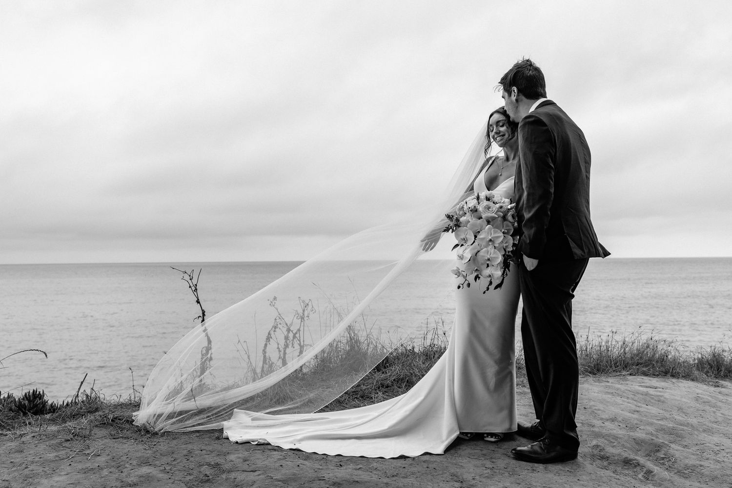 A wedding couple shares an intimate moment on a windswept coastal overlook.