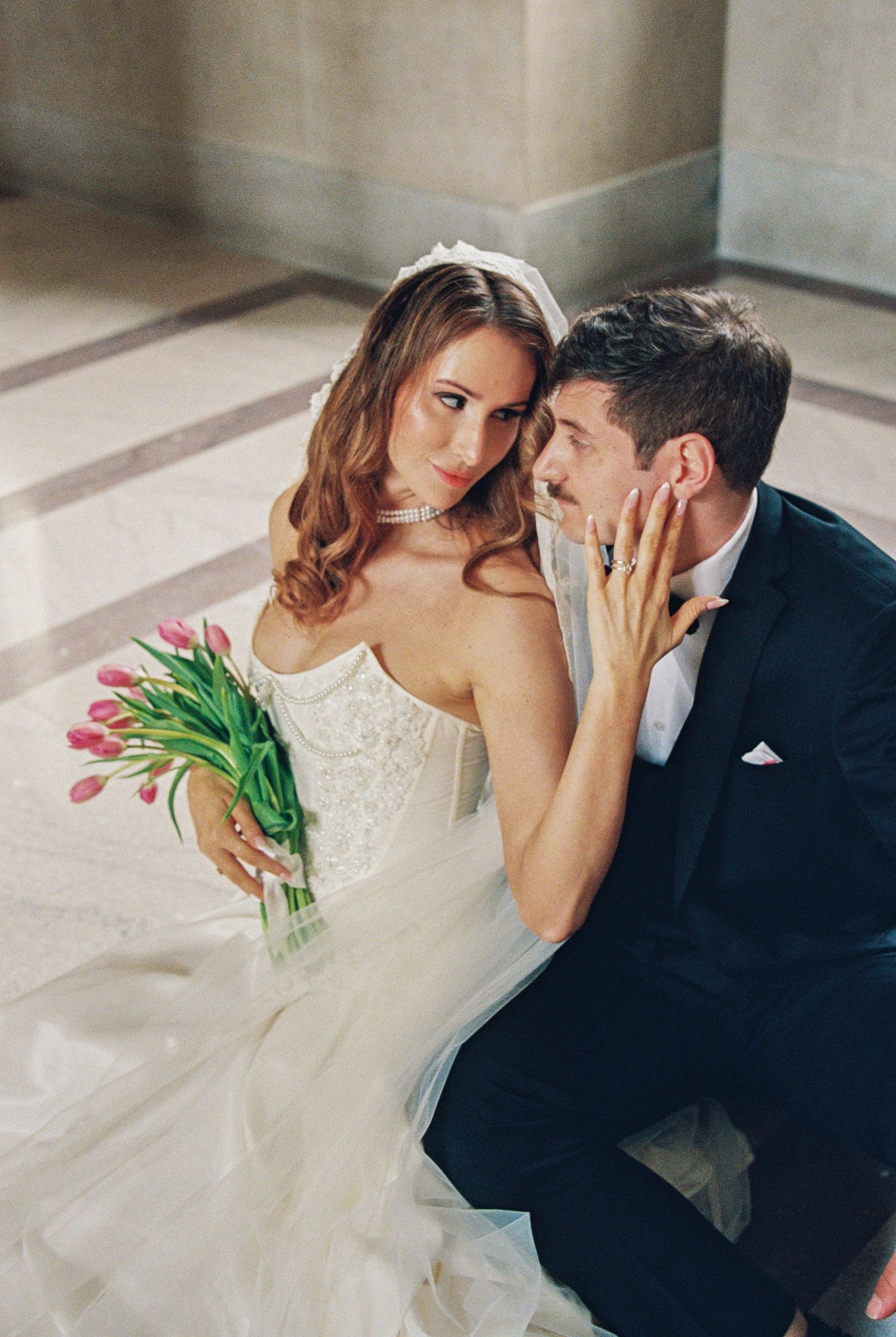 Bride in white lace gown holding pink tulips sits with groom in black tuxedo in elegant venue.