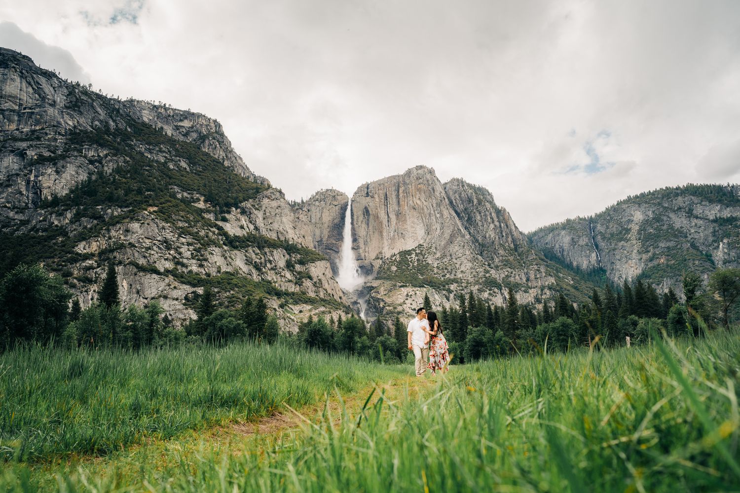 Couple standing in lush green meadow with Yosemite Falls and granite cliffs in background.