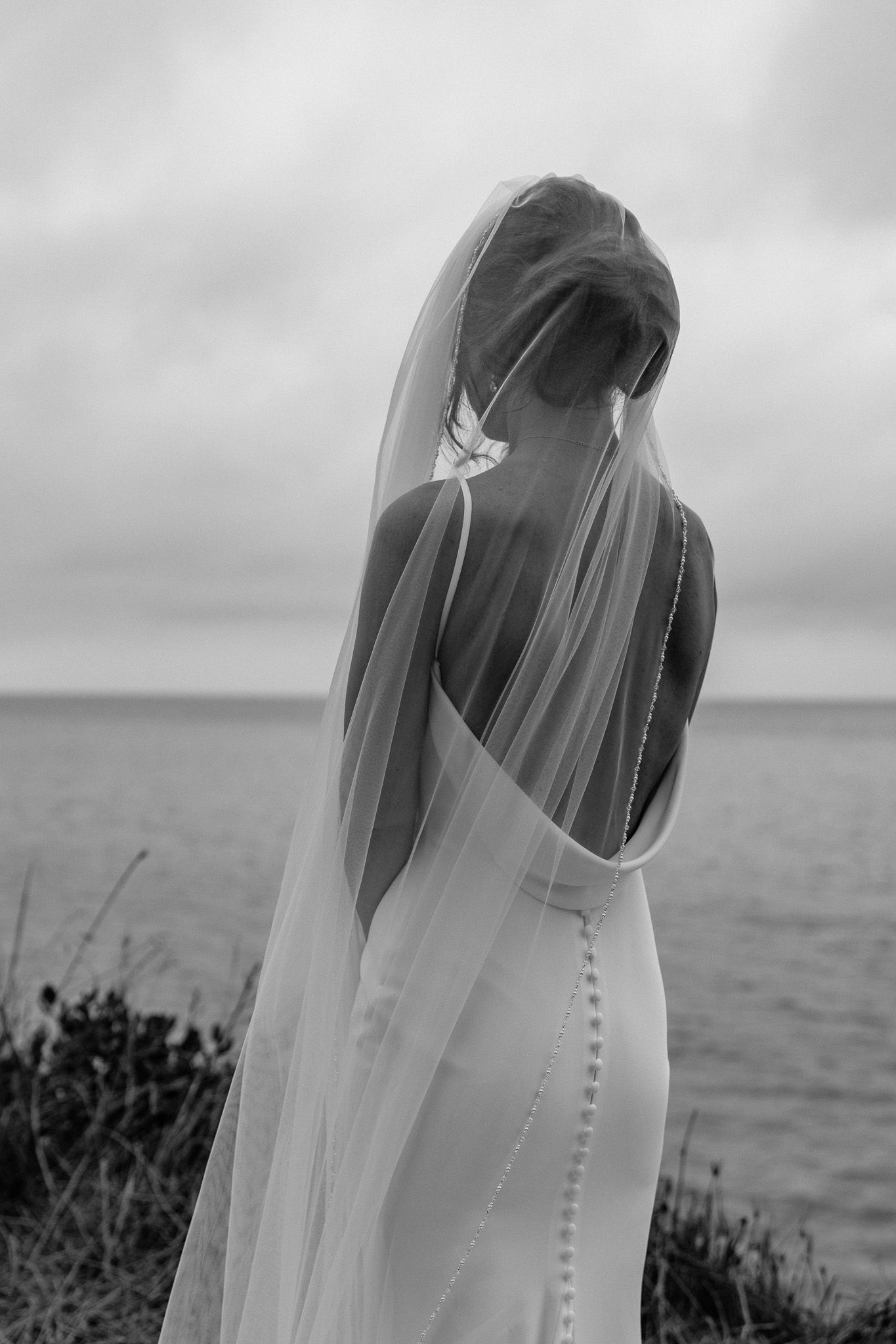 A flowing white wedding veil blows in the ocean breeze against a cloudy coastal backdrop.