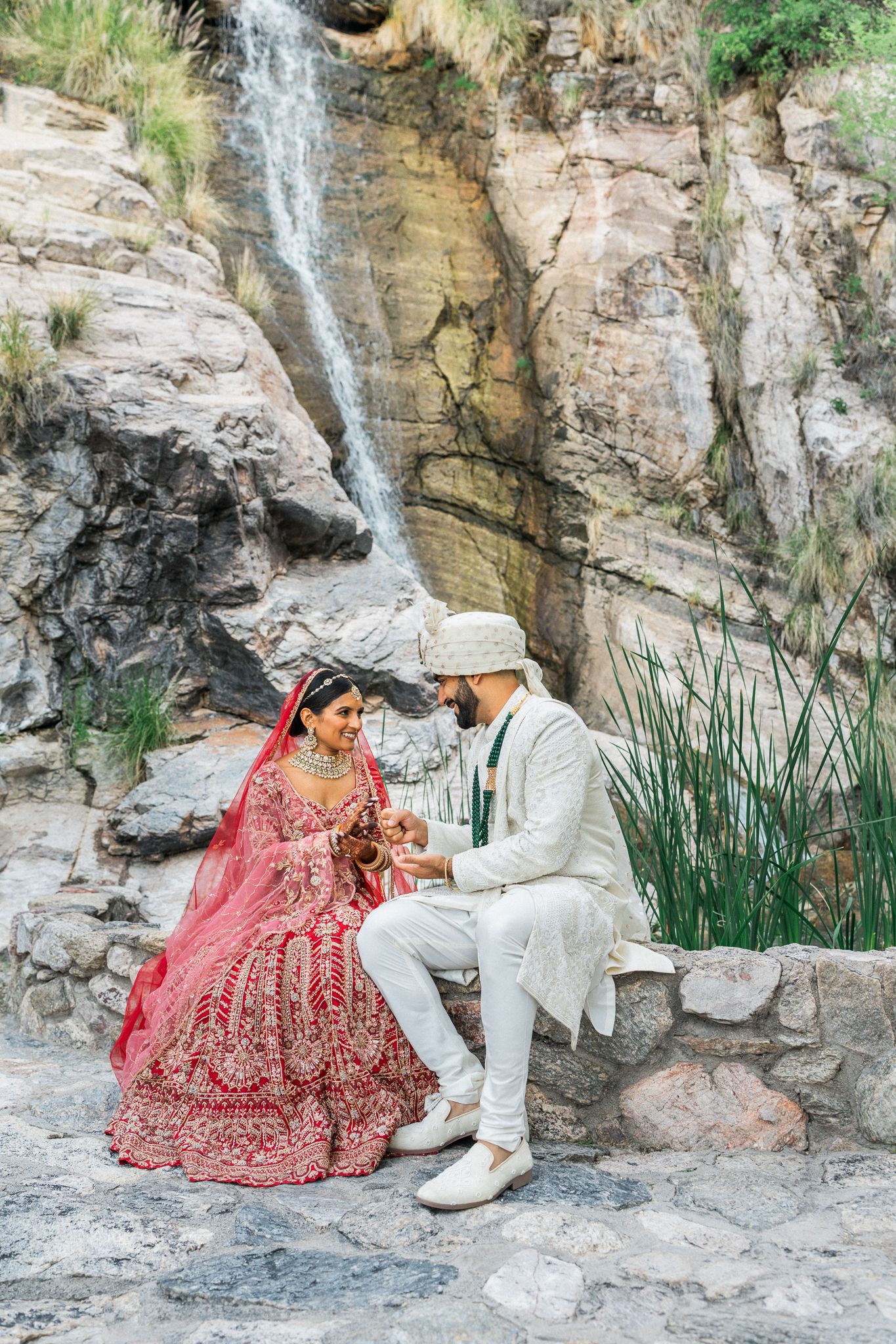 Wedding couple in traditional attire posing by natural waterfall and rock formation.