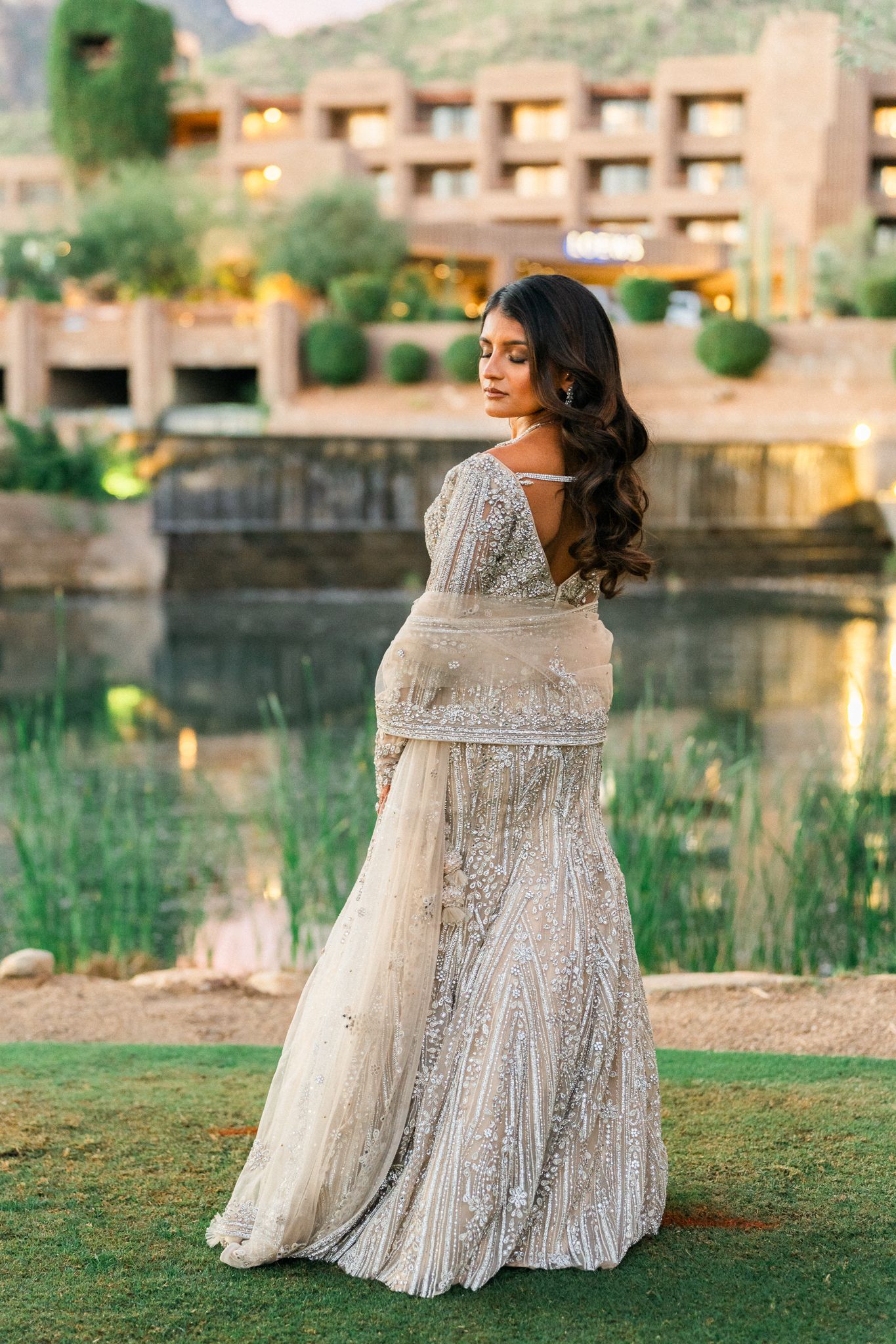A person in an embellished silver gown poses on grass near a reflective pond at dusk.