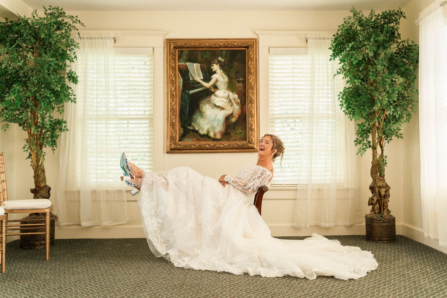 Bride in white gown sits elegantly, showing blue heels, with a classic portrait and potted trees in background.