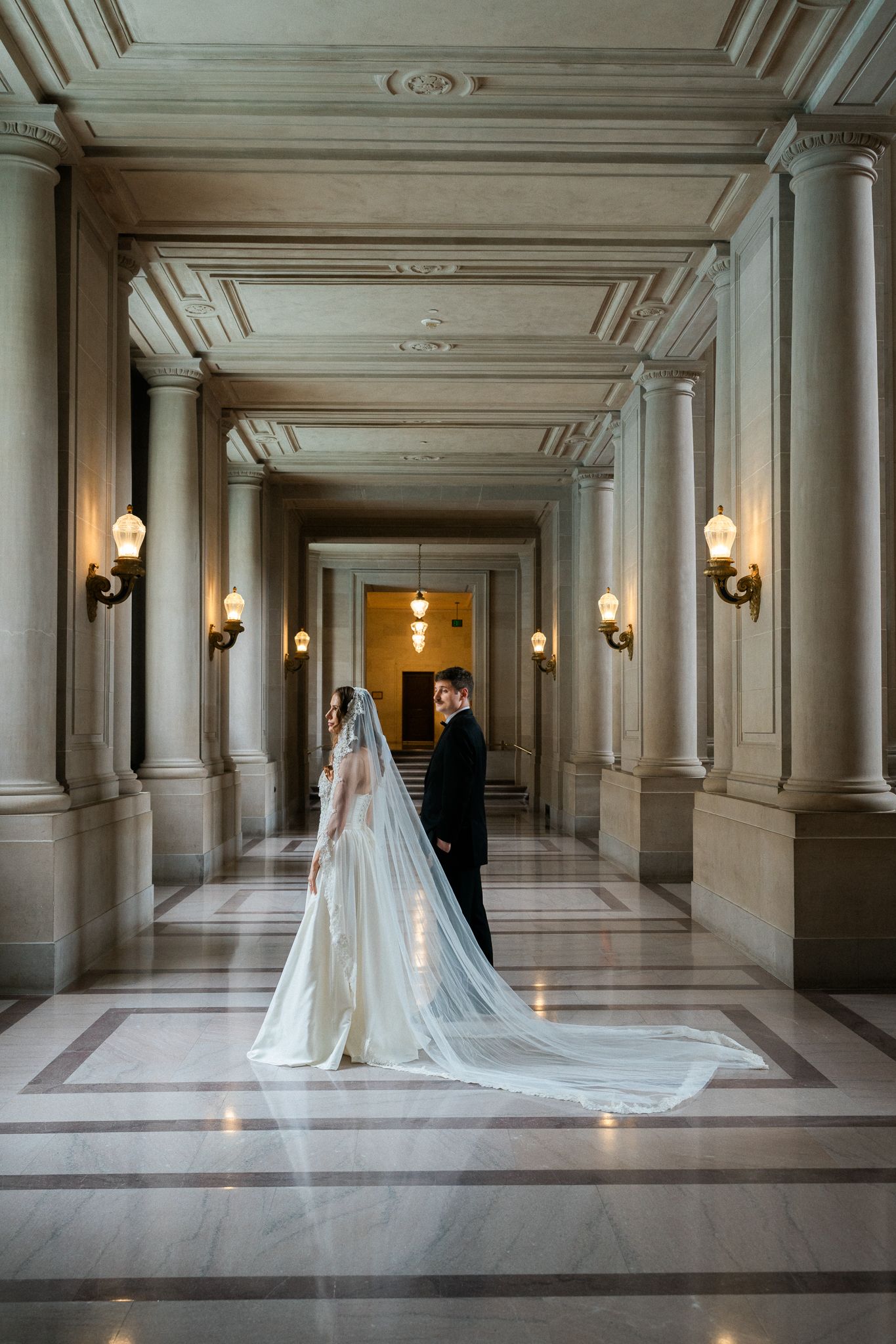 Bride and groom stand facing each other in an elegant marble hallway with tall columns and wall sconces.