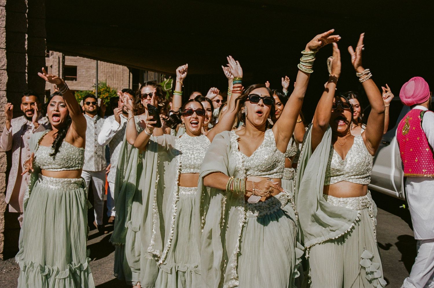 A group of bridesmaids in matching sage green outfits celebrate joyfully with raised arms at an outdoor wedding.