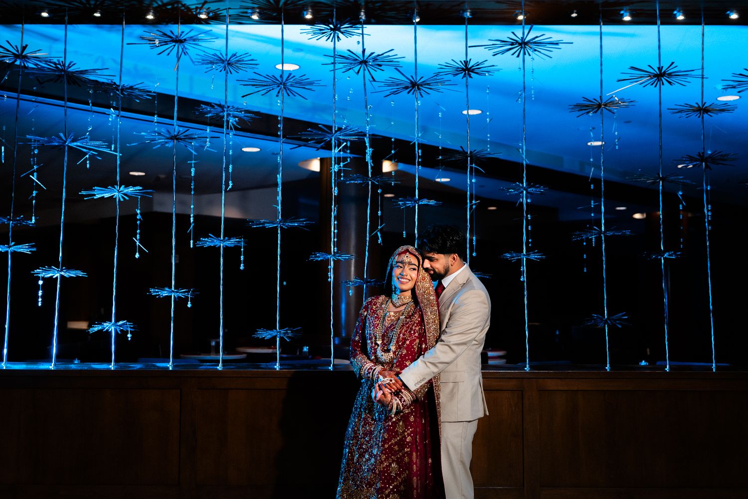 Couple in elegant wedding attire poses romantically under blue-lit decorative ceiling installation.