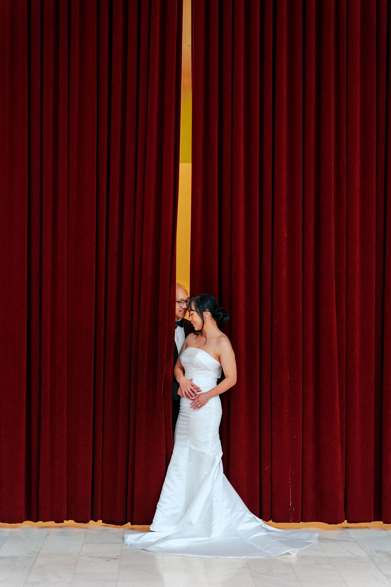 Bride in white gown kissing groom between red velvet curtains on a theater stage.